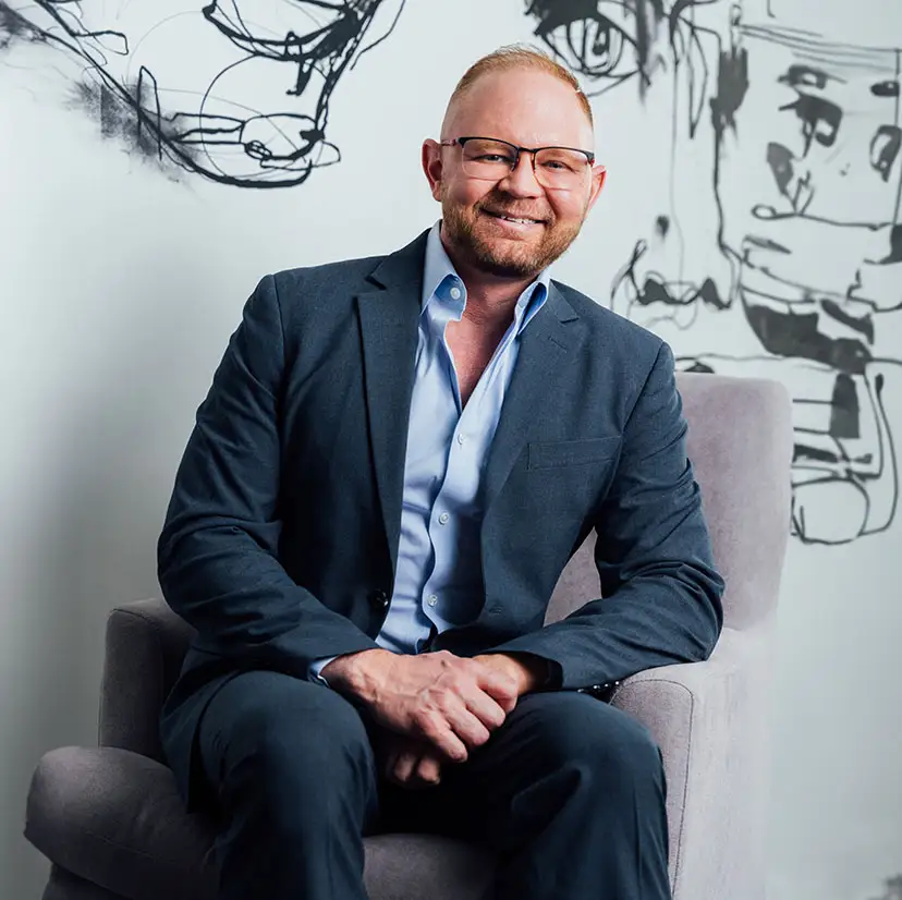 A portait shot of Dr. Craft in a dark blue suit and light blue under shirt sitting in a light grey chair in front of a black and white sketch wall paper. - in Phoenix, AZ
