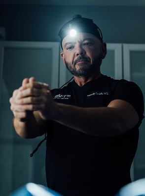 A dramatically lit, close-up photograph of Dr. Craft shows him in a surgical or medical setting. He is looking down intently with a serious expression and is wearing a headlamp with a bright, focused light illuminating his face from above. He is wearing a dark scrub top with his name, 'Rodger Craft, MD', visible on the chest. His hands are clasped and held up in front of him, possibly preparing for a procedure or demonstrating a technique. The background is dim, emphasizing his focused intensity.