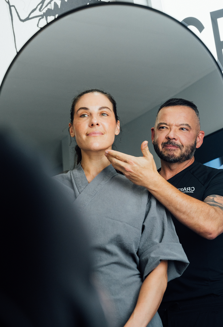A woman in a gray robe is smiling and looking off-camera. Standing behind her and to the right is a male plastic surgeon, Dr. Craft, with a beard and a black shirt, also smiling and pointing to the side of her face. The image appears to be taken in a clinical setting with bright lighting.