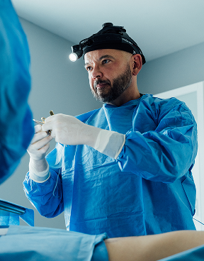 Dr. Randall Craft seen wearing blue protective scrubs and white latex gloves, prepping for surgery. He wears light head gear and a assistant is seen slightly on the left.