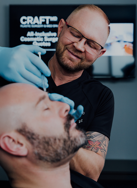 A focused, close-up photograph shows Dr. Matt Anderson (right) wearing glasses and blue gloves, smiling down as he carefully administers an injectable treatment to a male patient's face. The patient, who is bald and bearded, has his eyes closed. Dr. Anderson is wearing a dark shirt with the CRAFT MD logo visible on the collar. In the blurred background, a television screen displays the text "CRAFT MD Plastic Surgery & Med Spa".