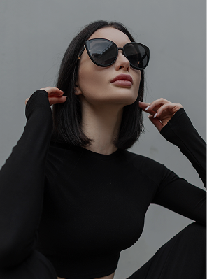 A low-angle, medium close-up photograph of a woman with short, dark hair, wearing large black sunglasses and a long-sleeved black top. She is looking up and away from the camera, touching the sides of her head with her hands, highlighting her facial structure and full lips. The background is a solid, light gray wall.