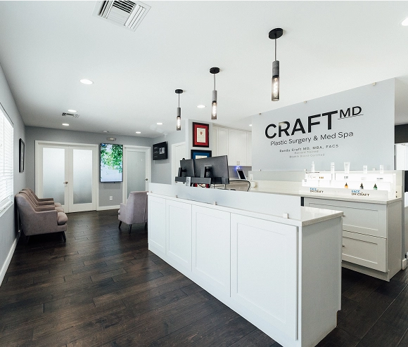 A wide interior photograph of the reception area and waiting room of a modern plastic surgery and med spa office. The prominent feature is the white reception desk with a sign that reads "CRAFT MD Plastic Surgery & Med Spa". The room has dark hardwood floors, light gray walls, and three pendant light fixtures over the desk. To the left is the waiting area with two plush, light-colored armchairs, a large vertical screen displaying an outdoor scene, and a set of double glass doors.
