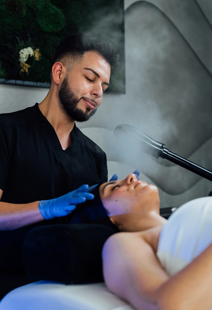 A photograph of an aesthetician, a man with a dark beard and hair, wearing black scrubs and blue gloves, gently touching the forehead of a female patient who is lying down with her eyes closed. The patient is receiving a facial treatment with a steamer positioned to the right, releasing a light mist. The background features a textured gray wall and a piece of abstract moss or preserved plant art.