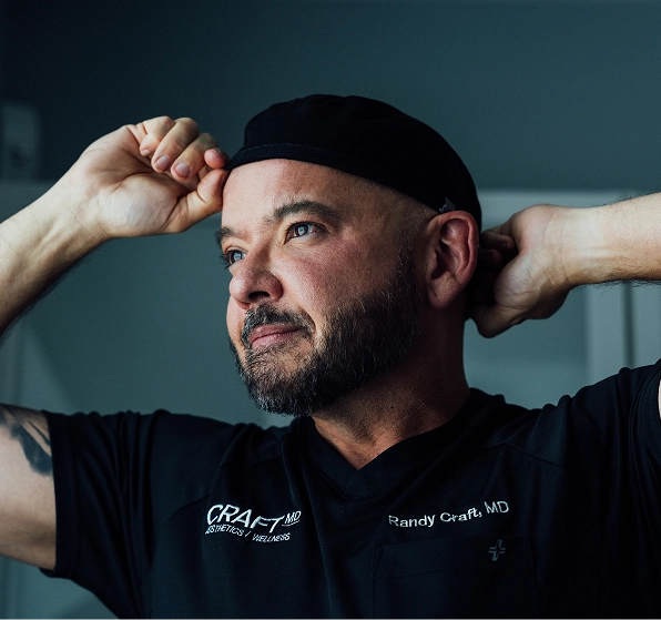 A focused, close-up portrait of Dr. Randy Craft shows him in profile, looking up and to the right with a determined expression. He is preparing for a procedure, pulling a black scrub cap over his head with both hands. He is wearing a black scrub top with the "CRAFT MD Aesthetics / Wellness" logo and his name, "Randy Craft, MD", embroidered on the chest. The background is a dim, uniform gray-blue.