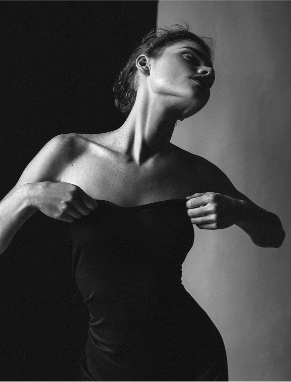 A high-contrast, black-and-white portrait of a woman wearing a strapless dark top, pulling up the fabric with both hands. She is looking dramatically up and to the right, highlighting the lean line of her neck and jaw. The background is split into two distinct, sharply defined vertical sections of black and light gray, creating a dramatic shadow effect on her figure.