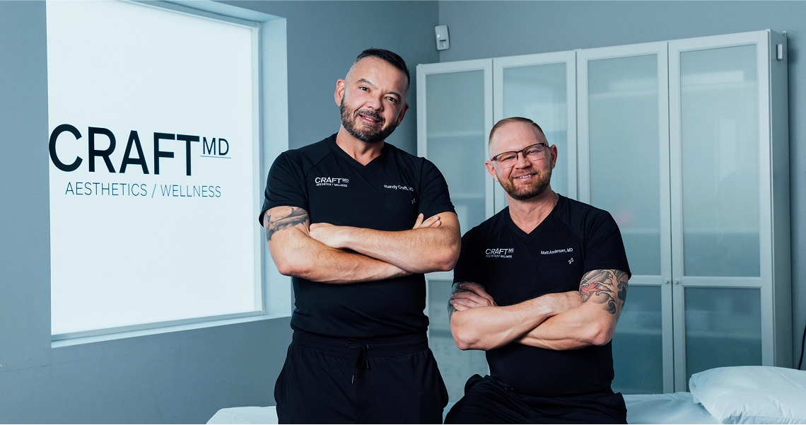 A photograph of two doctors, Dr. Randy Craft (left) and Dr. Matt Anderson (right), standing side-by-side in what appears to be a consultation or treatment room. Both men are smiling, have their arms crossed, and are wearing black scrubs embroidered with the "CRAFT MD" logo and their respective names. Behind them is a large, illuminated sign in a window that reads "CRAFT MD AESTHETICS / WELLNESS", and a wall of white glass-front cabinets.
