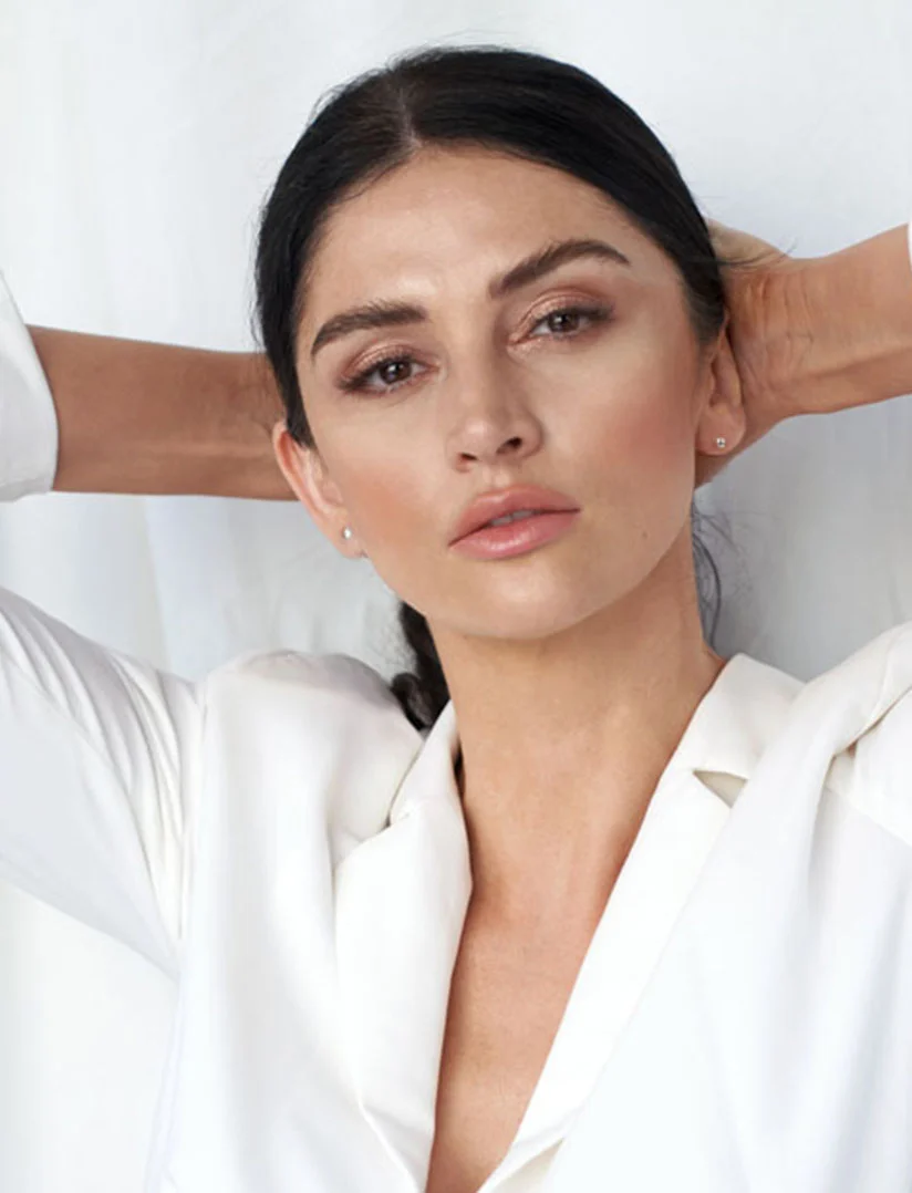 A close up of a confident woman with shoulder-length dark hair, styled with loose waves, poses against a backdrop of softly draped white fabric. She is wearing a white blazer over a simple white top. Her arms are raised, with her hands gently resting behind her head, tilting her chin up slightly as she looks directly at the camera with a self-assured expression. The lighting is soft and diffused, highlighting her features and the textures of the fabric. - Ultrasonic Guided Filler Injection Reversal in Phoenix, AZ