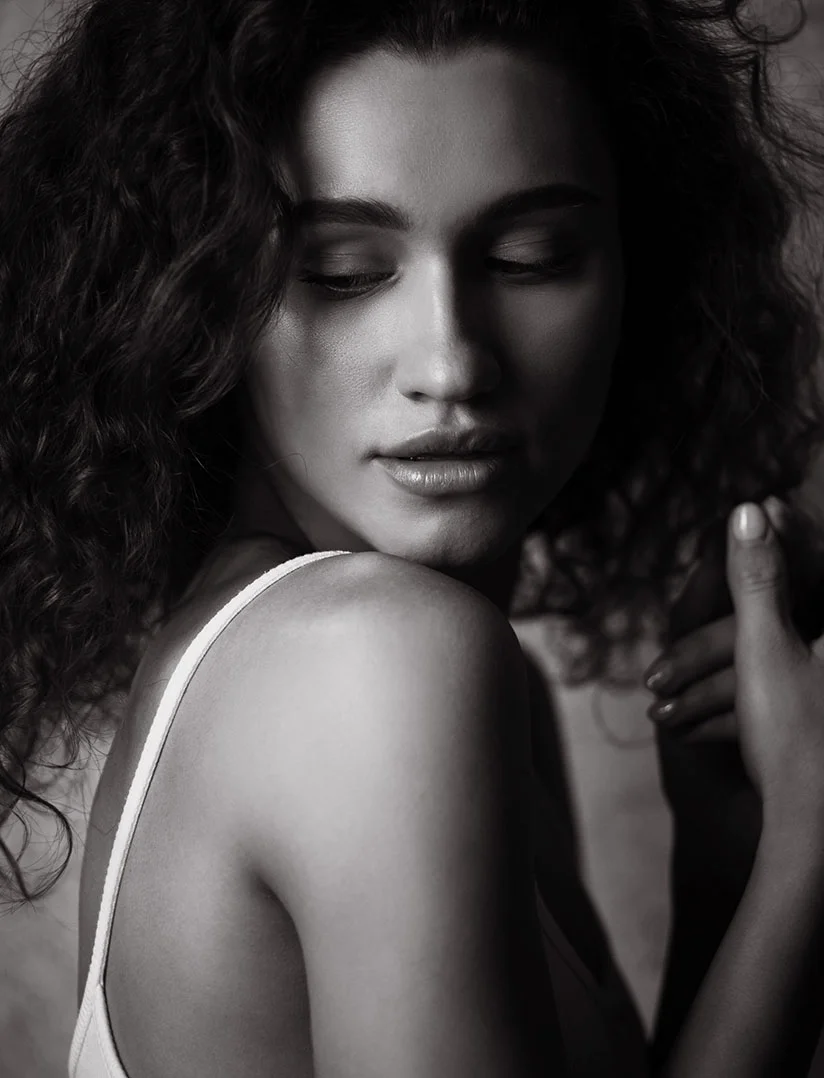 A high-contrast, black and white close-up of a young woman with long blonde hair, looking intensely at the camera. Her hands are clasped and resting on a dark surface against a solid dark background. - Arm Lift in Phoenix, AZ