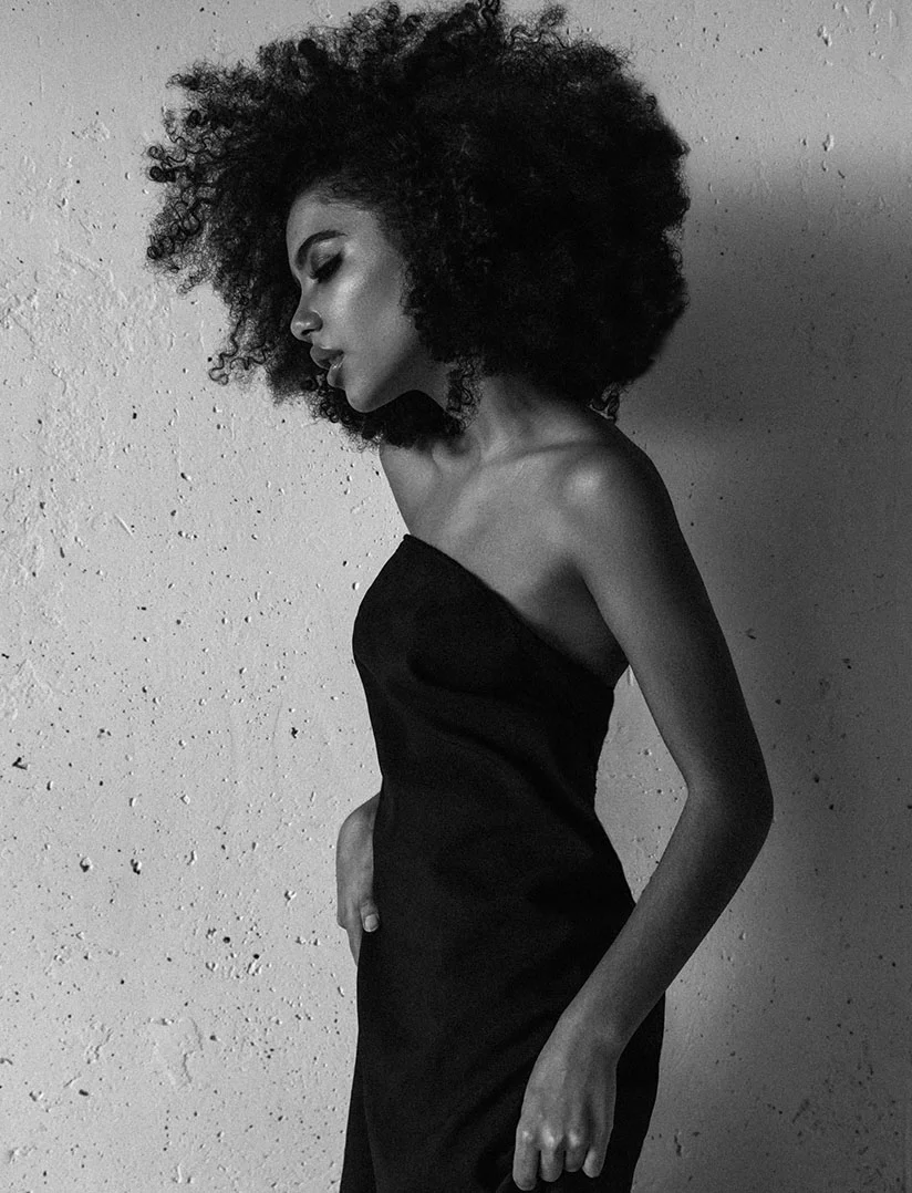A black and white portrait shot of a woman with large black hair standing sideways in front of a textured wall. She wears a strapless black dress as she glances down at the floor. - Botox and Wrinkle Relaxers in Phoenix, AZ