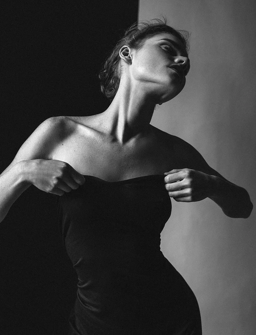A dramatic, high-contrast, black and white, vertical photo of a woman in a strapless black garment, pulling the material down slightly with her hands. Her head is tilted back, emphasizing her jawline and neck, and she is looking upward. The background is split between a deep black on the left and a light gray on the right. - Breast Fat Grafting in Phoenix, AZ