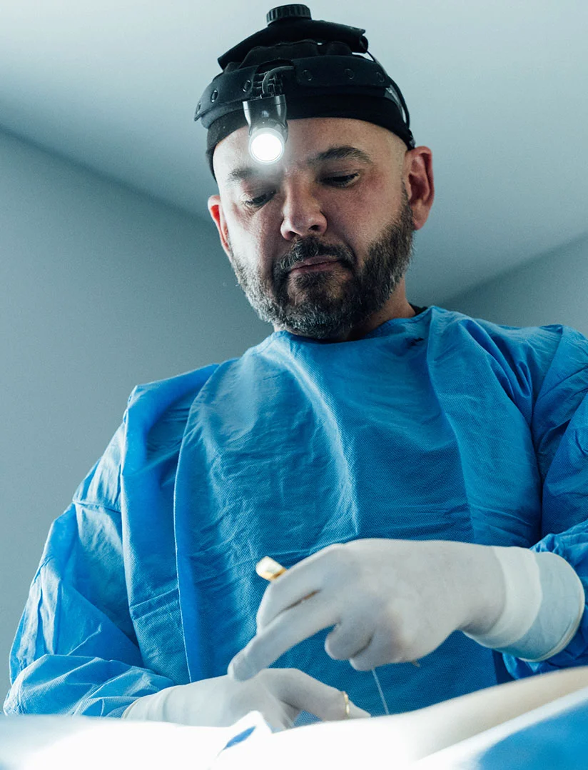 A vertical close-up photo of a male surgeon, Dr. Craft, in blue surgical scrubs, white gloves, and a surgical headlight. He is focused on a procedure, holding a fine suture or tool between his fingers. The scene is brightly lit. - Breast Implant Removal in Phoenix, AZ