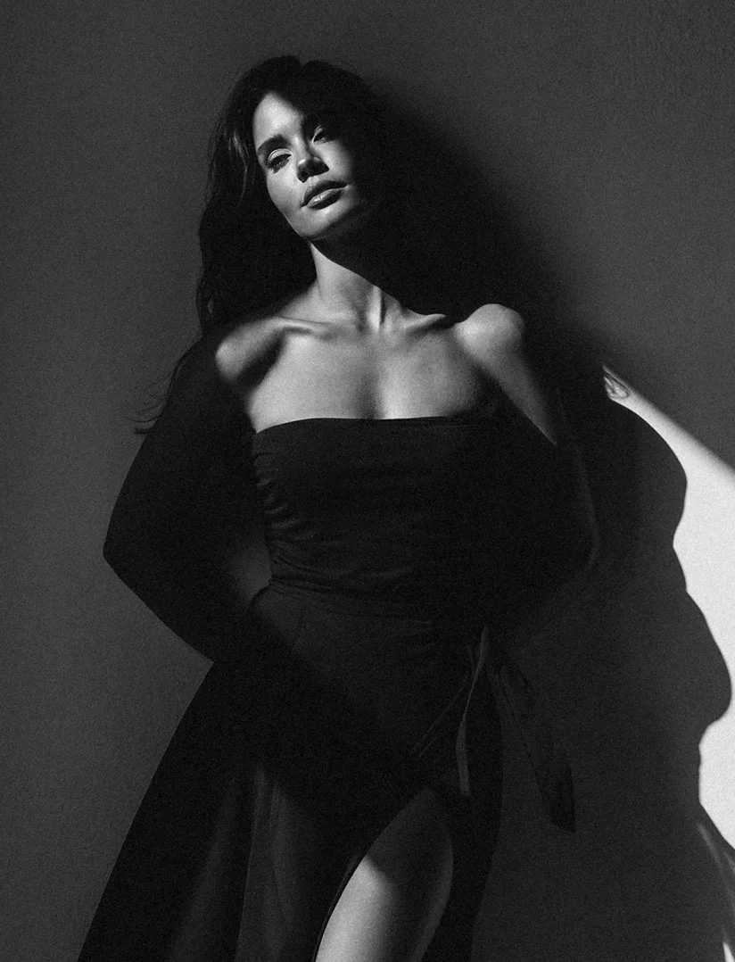 A dramatic, high-contrast, black and white, medium shot of a woman posing against a wall. She is wearing a strapless black dress with a high slit. Her face is partially illuminated by a strong, directional light, casting deep shadows on the wall and emphasizing her neck, shoulders, and clavicle. She is looking directly at the camera with an intense expression. - Breast Reduction in Phoenix, AZ