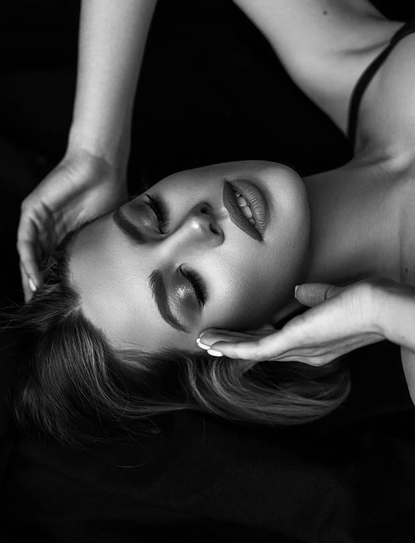 A dramatic, vertical, high-contrast, black and white close-up photo of a woman's face lying down with her eyes closed, suggesting a state of rest or treatment. Two hands, with light-colored fingernails, are gently cupping her face near her jawline and temples. The lighting creates deep shadows and highlights on her cheekbones and lips, with a solid black background. - Brow Lift in Phoenix, AZ