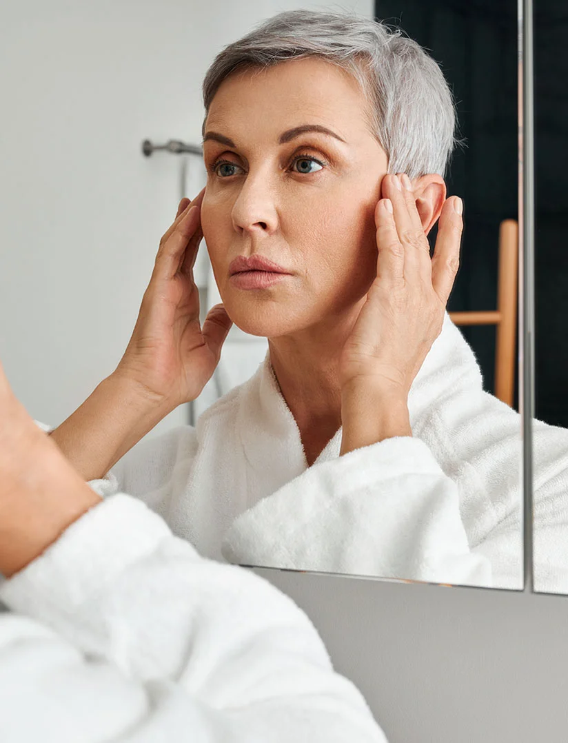A vertical photo of an older woman with short gray hair wearing a white terrycloth bathrobe, looking intensely at her reflection in a bathroom mirror. She is gently touching her temples and cheeks with both hands, as if evaluating her skin. - Croton Oil Peel in Phoenix, AZ