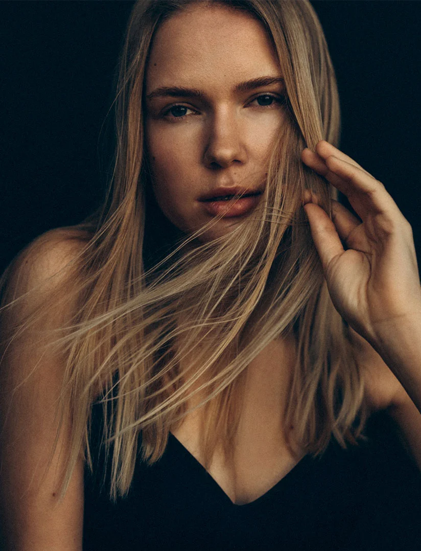 A dramatic, vertical, close-up photo of a young woman with long blonde hair. She is looking directly at the camera with a calm, serious expression, and her left hand is raised near her temple, brushing back a few strands of hair. She is wearing a dark, low-cut top. The background is a solid, deep black, creating a high-contrast and intimate portrait. - Dermal Fillers in Phoenix, AZ