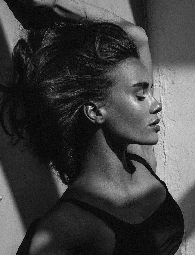 A dramatic, high-contrast, black and white profile photograph of a woman with her eyes closed, leaning her head against a textured wall. She is wearing a dark tank top, and her hair is pulled up. Sharp, diagonal shadows cut across her face, neck, and chest, emphasizing her bone structure. - Ear Surgery in Phoenix, AZ