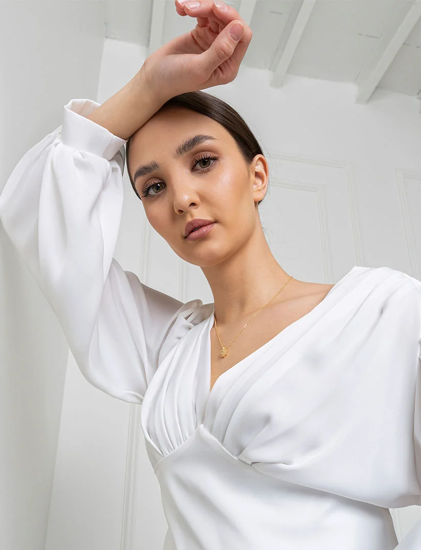 A high-angle, close-up photograph of a woman with dark hair pulled back, looking directly at the camera. She is wearing a white dress or top with voluminous, cuffed sleeves and a gold pendant necklace. One arm is raised, with her hand resting on her forehead. The background is a bright white room with visible ceiling beams. - Eyelid Surgery in Phoenix, AZ