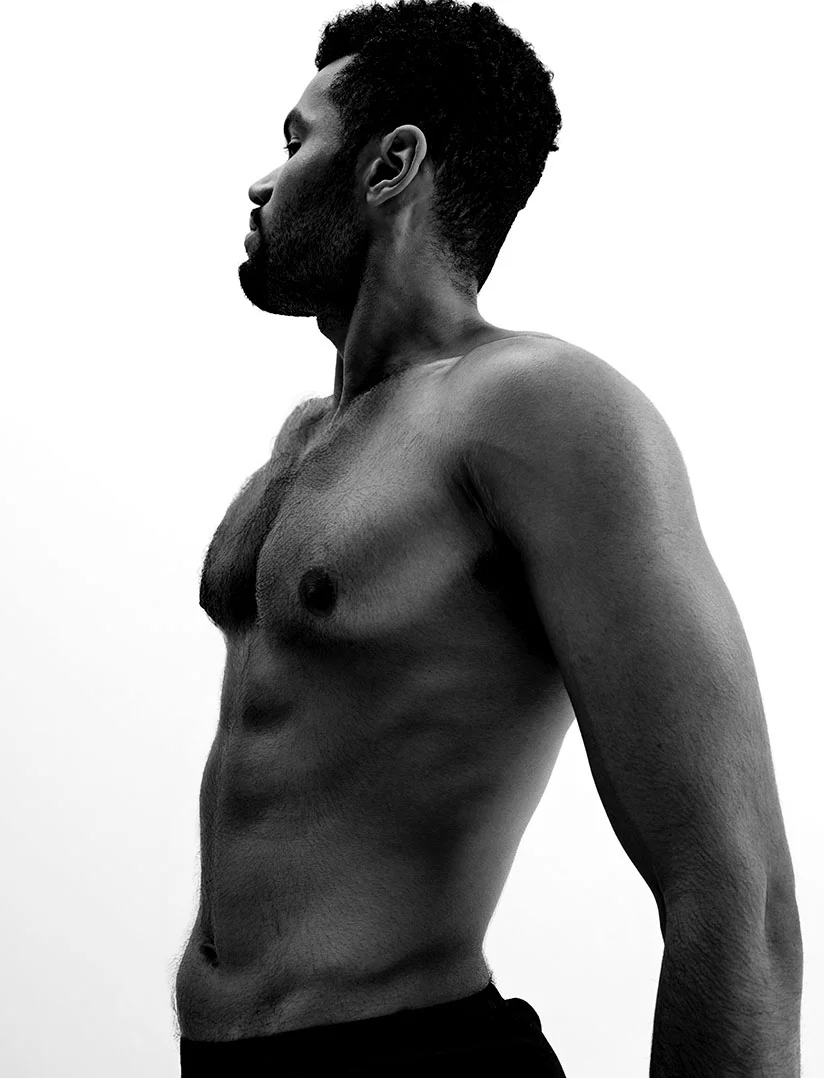 A stark, high-contrast, black and white profile photograph of a shirtless man with a dark, trimmed beard and short, curly hair. He is standing against a bright white background, showing his muscular torso and strong profile, looking to his left. - Face Body Implants for Men in Phoenix, AZ