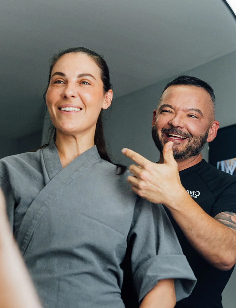 A woman in a gray robe is smiling and looking off-camera. Standing behind her and to the right is a male plastic surgeon, Dr. Craft, with a beard and a black shirt, also smiling and pointing to the side of her face. The image appears to be taken in a clinical setting with bright lighting. - Face Lift in Phoenix, AZ