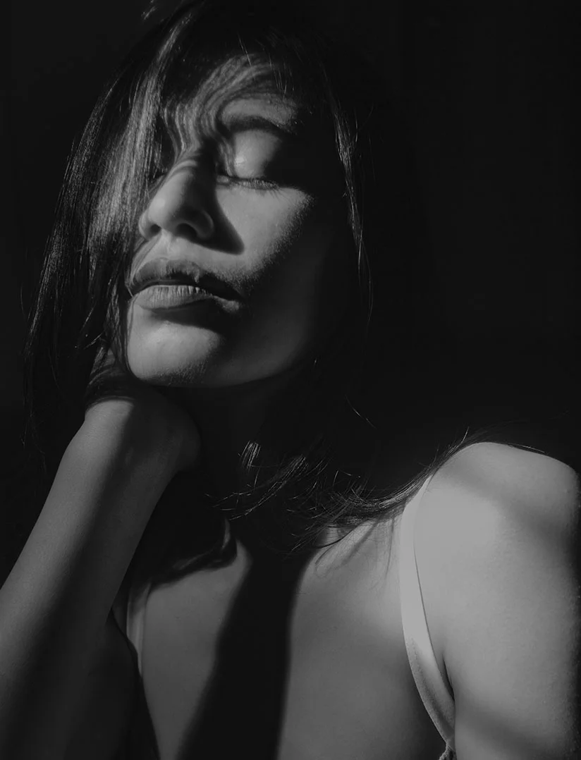 A highly dramatic, vertical, black and white close-up photo of a woman with long dark hair, tilted back with her eyes closed. Her left hand is resting around the back of her neck. A single, strong light source illuminates her face and upper chest, emphasizing her lips and jawline while creating deep shadows in the background and over part of her face. - Facial Neck Liposuction in Phoenix, AZ