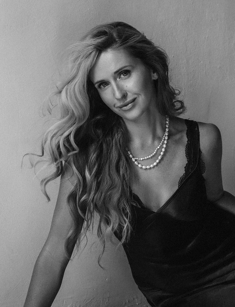 A black and white, vertical, medium close-up photo of a woman with long, wavy blonde hair, looking directly at the camera with a slight smile. She is wearing a dark, low-cut slip dress with lace detail and a double strand of pearls. Her hair is cascading over her shoulders and partially across the frame. She is posed against a plain, light-colored wall. - Facial Rejuvenation in Phoenix, AZ