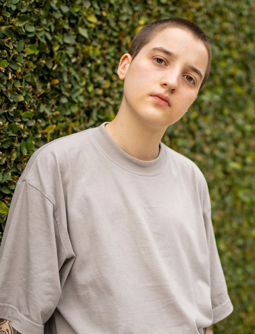 A horizontal photo of a young person, who appears male, with dark hair, resting an elbow on a cushion and looking contemplatively out a window. The person is wearing a black t-shirt. Window blinds create horizontal light and shadow lines across the background. - Female to Male in Phoenix, AZ