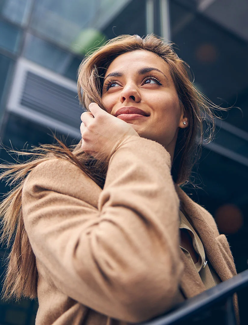A low-angle, medium close-up photo of a young woman with long, flowing brown hair, wearing a beige coat. She is looking up and slightly to her left with a thoughtful, happy expression, with one hand resting near her chin. The background is a modern building with dark, reflective glass and geometric lines. - Hair Restoration in Phoenix, AZ