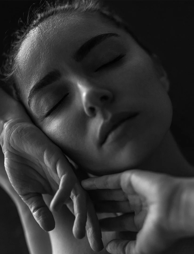 A dark, high-contrast, black and white close-up photo of a woman's face with her eyes closed, suggesting deep relaxation or repose. Two hands gently cup or cradle her face and chin. The lighting is focused on her face, with a dark, blurred background. - Hydra facial in Phoenix, AZ