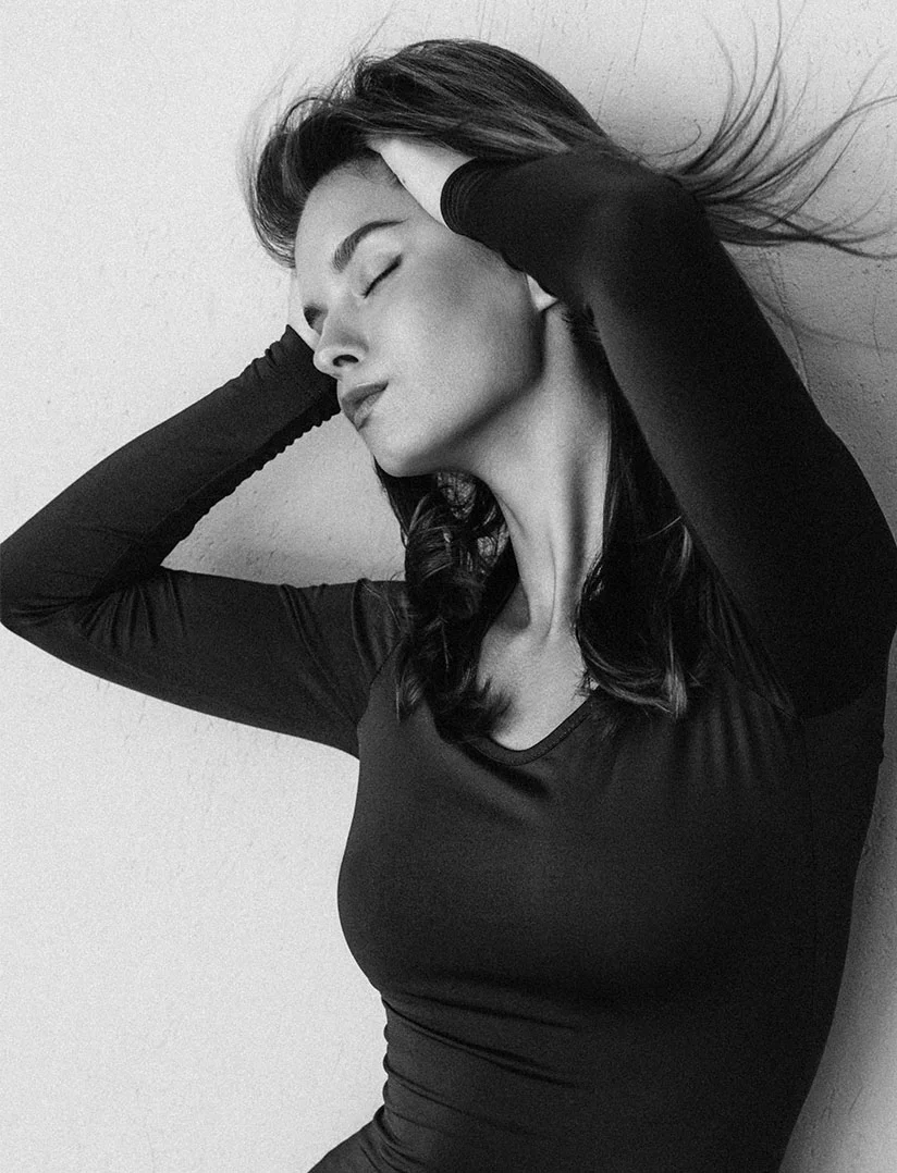 A close-up, black and white photograph of a woman with her eyes closed, pressing both hands to the sides of her head near her ears. She is wearing a tight, long-sleeved black top. Her hair is slightly windswept to the right. - Insta Lift in Phoenix, AZ
