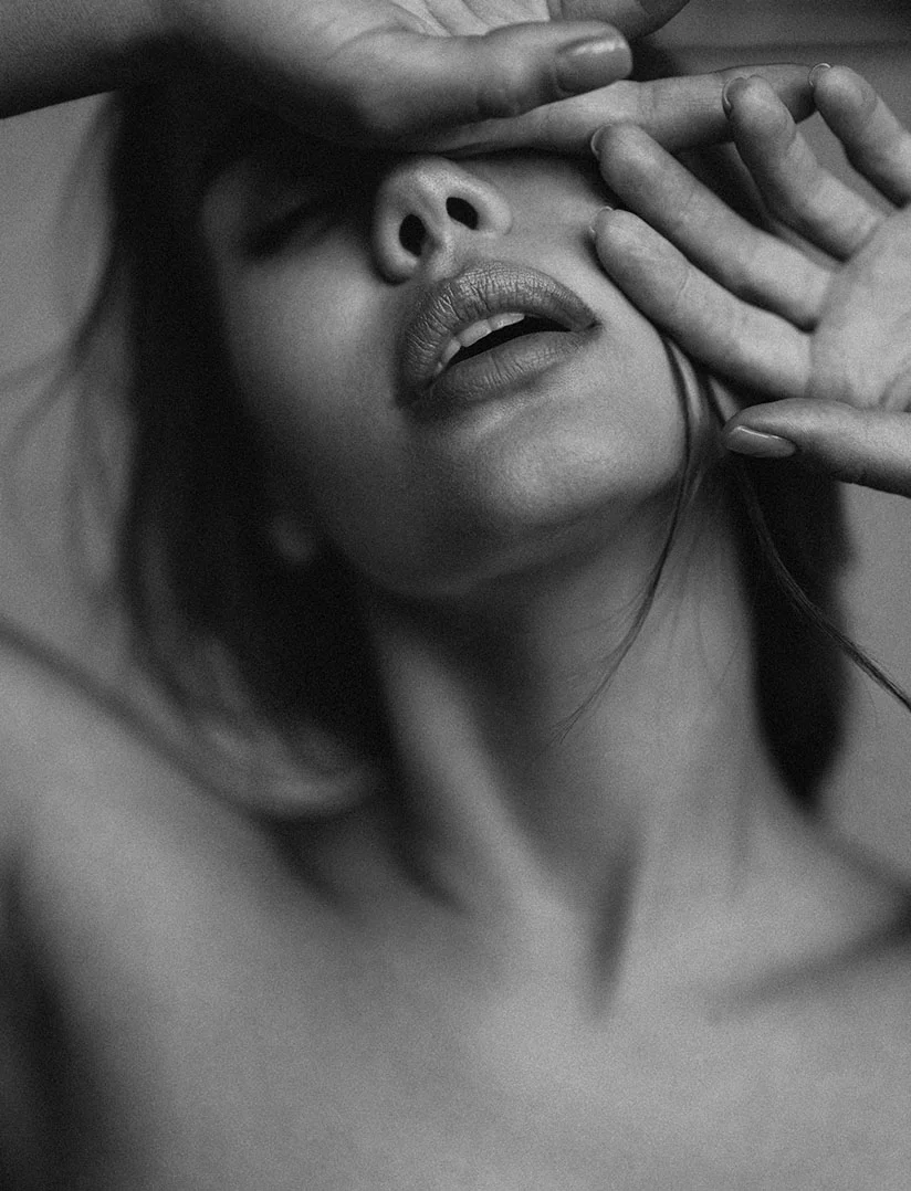 A high-contrast, black and white, close-up photo of a woman's head and neck tilted back, with her mouth slightly open. Both of her hands are covering her eyes and forehead. The image focuses on her expressive mouth and jawline, with dramatic shadows and highlights. - Lip Fillers in Phoenix, AZ