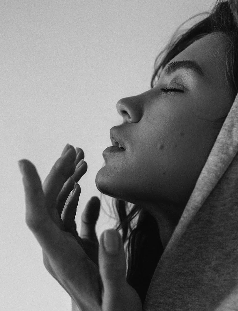 A striking, high-contrast, black and white close-up photo of a woman's profile with her eyes closed and her head tilted back, wearing a hooded garment that frames her face. Her mouth is slightly open. She is holding her hands up near her face, in an expressive or contemplative gesture. The background is a bright, plain white or light gray.A striking, high-contrast, black and white close-up photo of a woman's profile with her eyes closed and her head tilted back, wearing a hooded garment that frames her face. Her mouth is slightly open. She is holding her hands up near her face, in an expressive or contemplative gesture. The background is a bright, plain white or light gray. - Lip Lift in Phoenix, AZ