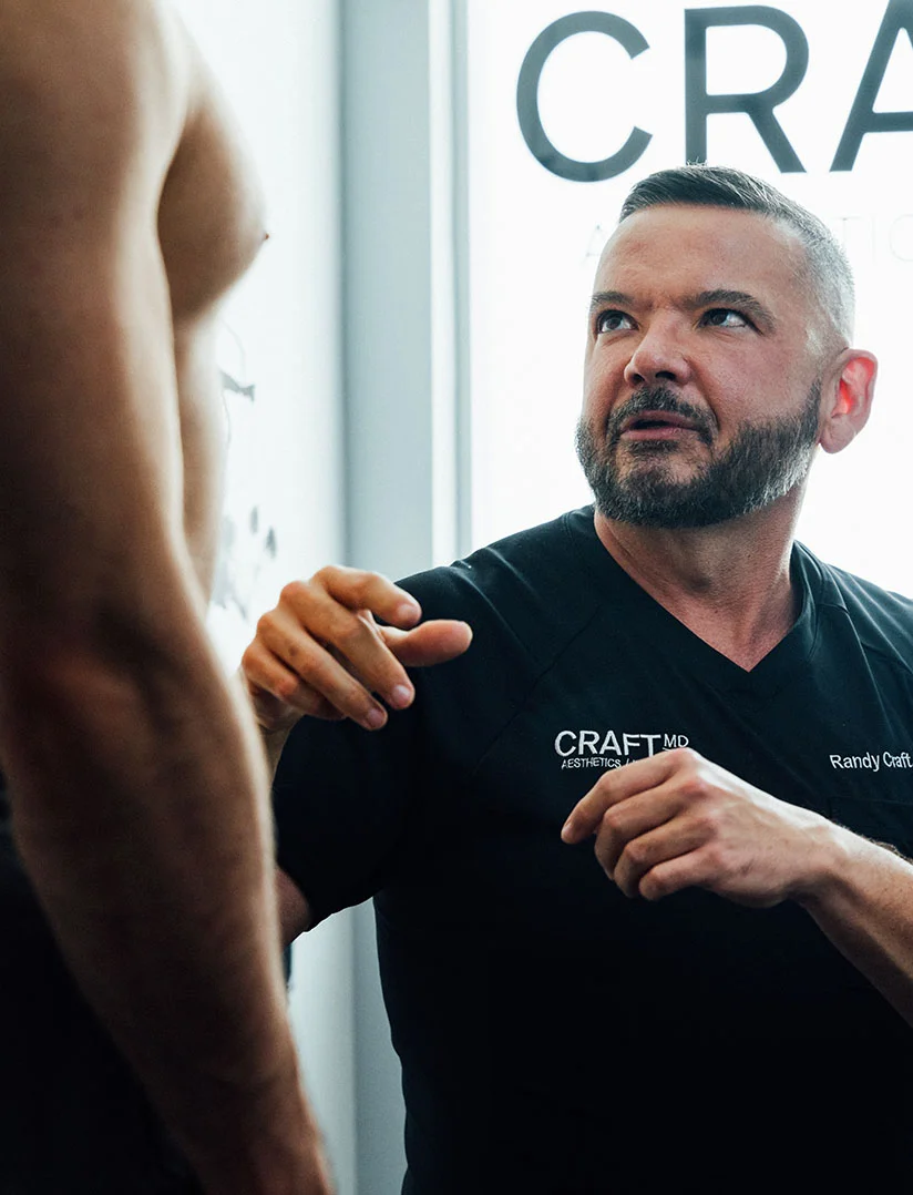 A medium close-up photo of Dr. Randy Craft in black scrubs, looking up and to the left with a focused expression while speaking to a tall, bare-chested male patient, whose shoulder and torso are visible in the foreground. Dr. Craft is resting his right hand on the patient's shoulder. The background features a large white sign with the word "CRAFT" partially visible. - Liposuction for Men in Phoenix, AZ