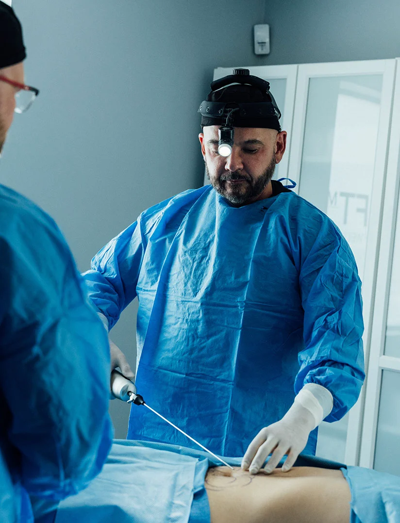 A vertical photo showing a surgeon, Dr. Craft, wearing a blue surgical gown, white gloves, and a headlamp, performing a liposuction procedure. He is holding a cannula-like instrument in his right hand and has his left hand resting on the patient's abdomen, which has surgical markings drawn on it. A second person's back is visible in the left foreground. - Liposuction in Phoenix, AZ