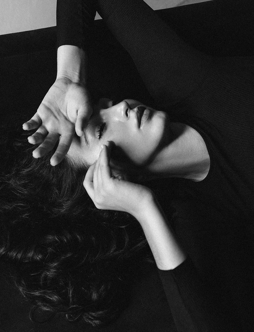 A high-contrast, black and white, close-up photo of a woman lying down, looking up with her eyes closed or partially closed. She is wearing a dark, long-sleeved shirt. One hand is covering her forehead and the other is gently touching her cheek, creating a dramatic, expressive pose. Her dark, curly hair frames the lower part of the image. - Liquid Rhinoplasty in Phoenix, AZ
