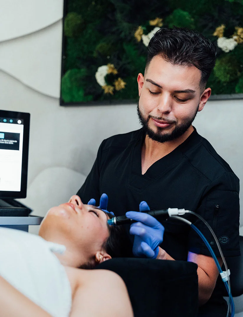 A close-up photograph of a male aesthetician with a beard and dark hair, wearing a black scrub top and blue gloves, performing a skin treatment on a female client. He is using a handheld device on the client's forehead. A computer monitor and a dark green, moss-like wall decoration are visible in the background. - Microneedling in Phoenix, AZ