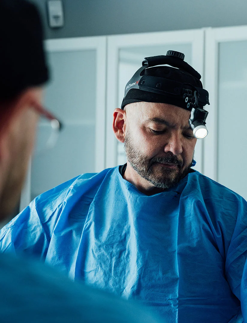 A portrait photo of a male surgeon, Dr. Craft, wearing a surgical headlamp and a blue surgical gown. He has a shaved head and a beard and is looking down and concentrating. A person in a blue scrub top and a surgical cap is partially visible in the blurred foreground, facing away from the camera. The background shows white, cabinet-like surfaces. - Mini Facelift in Phoenix, AZ