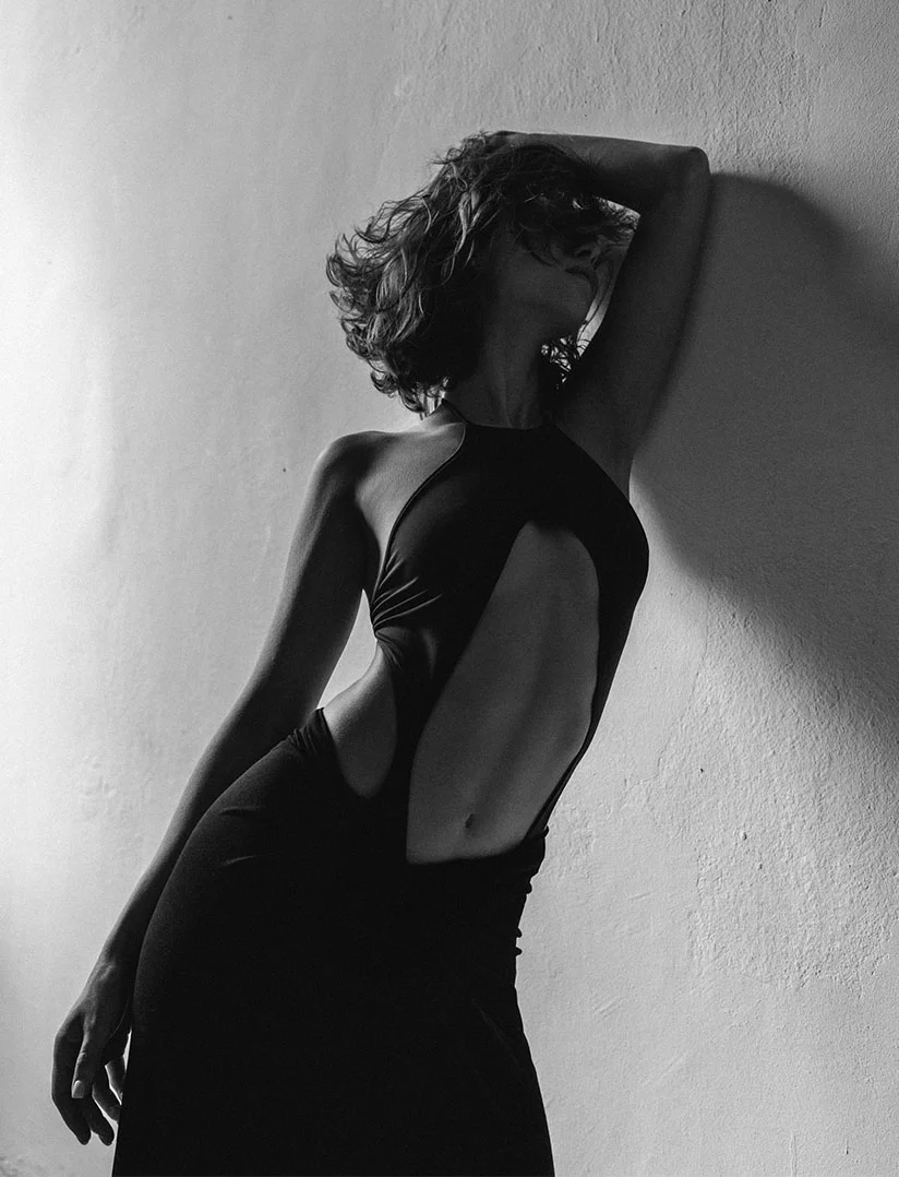A high-contrast, black and white photograph of a slender woman with curly hair leaning against a textured wall. She is wearing a dark halter dress with large side and torso cut-outs that expose her midriff. One arm is raised, with her hand resting on her head. - Mini Tummy Tuck in Phoenix, AZ