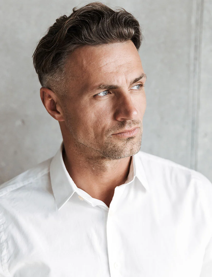 A close-up portrait of a middle-aged man looking intensely to the right. He has short, styled, dark hair, light eyes, and light stubble on his chin and jawline. He is wearing a crisp white collared shirt. The background is a slightly blurred, textured gray wall. - Minimally Invasive Neck Lift in Phoenix, AZ