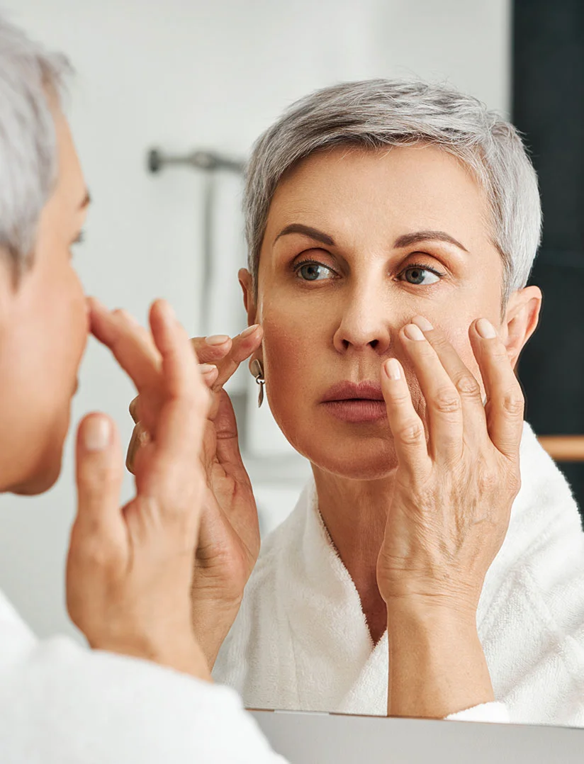 A close-up photo of a middle-aged woman with short, styled gray hair, wearing a white bathrobe, looking intently at her reflection in a bathroom mirror. She is touching her face near her cheek and eye area with the fingertips of both hands, with a contemplative or concerned expression. - MOHs Reconstruction in Phoenix, AZ