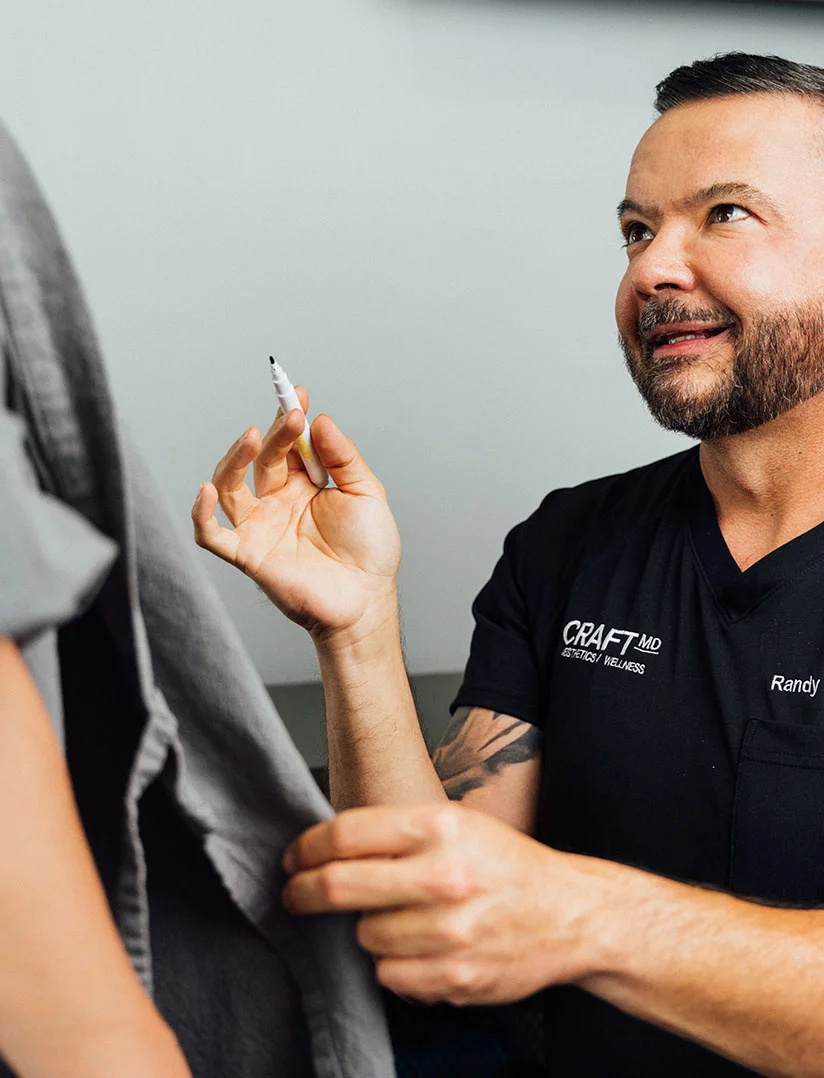 A close-up photo of Dr. Randy Craft in a black scrub shirt with the "CRAFT MD AESTHETICS / WELLNESS" logo, smiling and looking up and to the left at a patient, who is partially visible in the left foreground wearing a gray garment. Dr. Craft is holding a white marker or pen in his right hand, ready to make a mark. He has a beard and short, dark hair. - Mommy Makeover in Phoenix, AZ