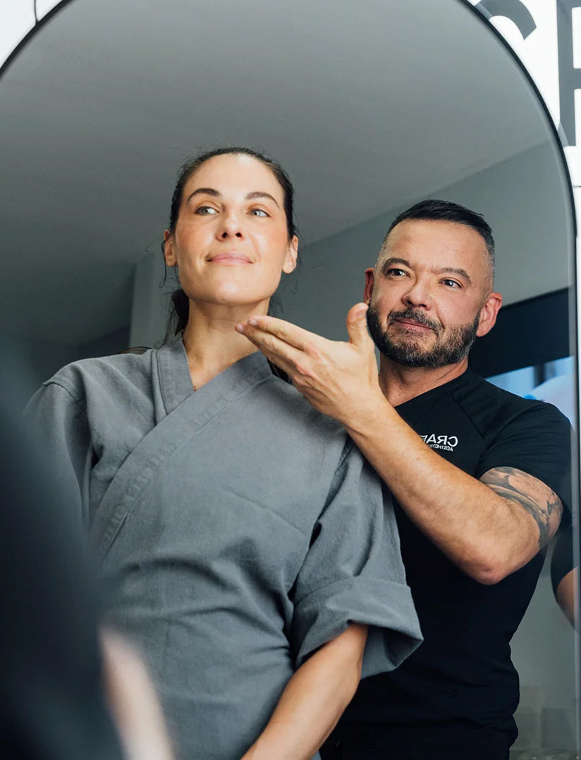 A woman in a gray robe is smiling and looking off-camera. Standing behind her and to the right is a male plastic surgeon, Dr. Craft, with a beard and a black shirt, also smiling and pointing to the side of her face. The image appears to be taken in a clinical setting with bright lighting. - Neck Lift in Phoenix, AZ