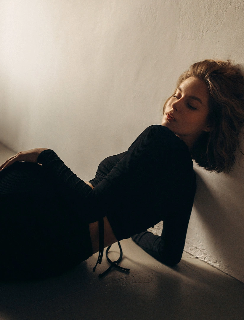 A dramatic photograph of a woman with short, wavy hair lying on her side on the floor, leaning against a white wall. She is wearing a long-sleeved black top that exposes a small strip of her midriff. Her eyes are closed, and the image is lit by a soft light from the upper right, casting a warm glow and subtle shadow. - Non-Surgical BBL in Phoenix, AZ
