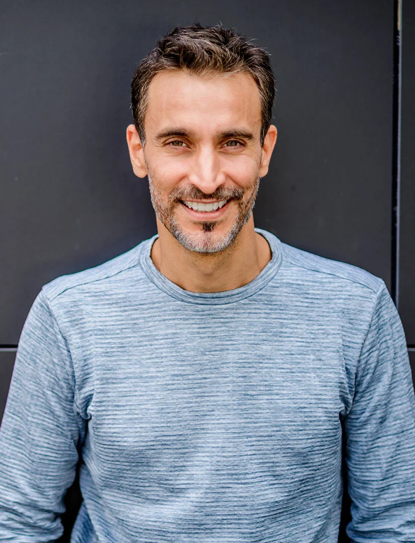 A smiling, handsome, middle-aged man with salt-and-pepper facial hair and dark hair is looking directly at the camera. He is wearing a long-sleeved, light blue, horizontally-textured shirt and is positioned against a dark gray or black wall. - P Shot in Phoenix, AZ
