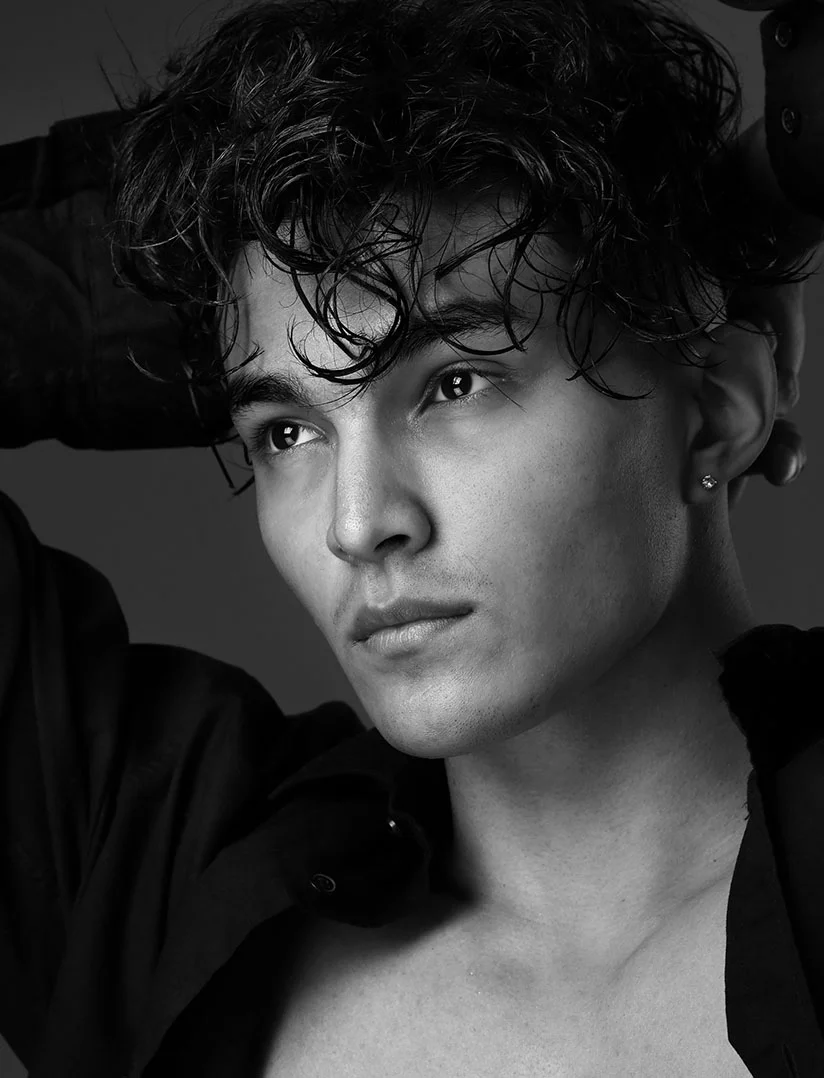 A high-contrast, black and white close-up portrait of a young man with dark, wet, curly hair. He is looking up and away from the camera, with one hand near his head. He wears an unbuttoned dark shirt and a small stud earring in his left ear. The background is a solid, dark gray. - Phalloplasty Filler in Phoenix, AZ