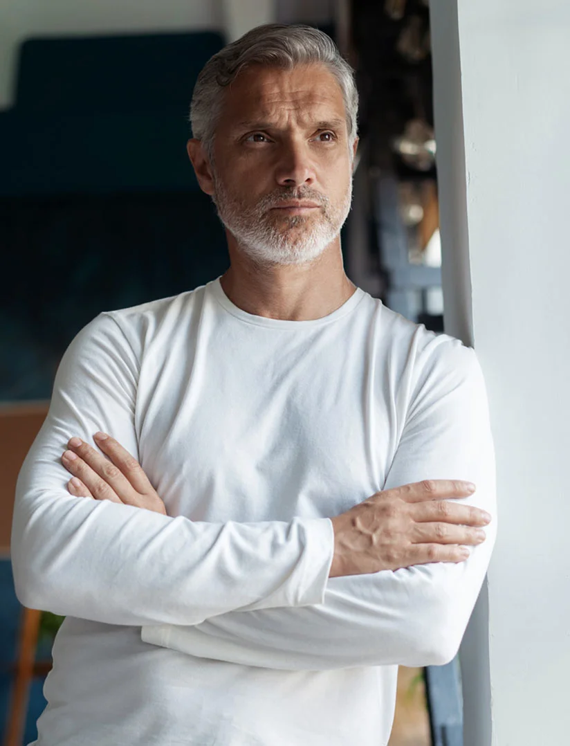 A portrait of a gray-haired man with short stubble, wearing a white long-sleeve shirt and leaning against a white wall with his arms crossed. He has a neutral expression, while the blurred background suggests a home interior with people conversing, a teal sofa, and a wooden chair with a blue cushion. The composition emphasizes the man as the central subject, framed against a softly out-of-focus setting. - PRF Hair Restoration in Phoenix, AZ