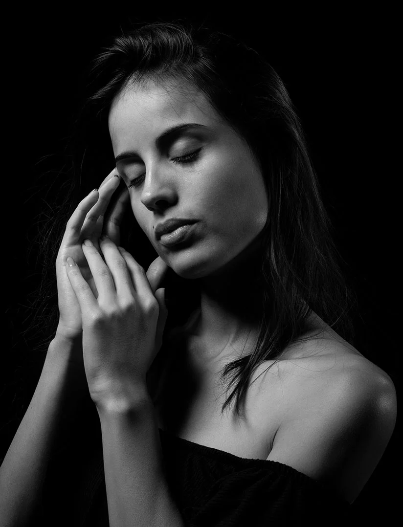 The image is a black-and-white portrait of a woman with shoulder-length dark hair, fair skin, and an off-the-shoulder black garment. Her right hand delicately touches her left ear as she looks downward, her left eye closed in a serene expression. The dark background, accented by white lighting highlights, creates a dramatic contrast that emphasizes her contemplative pose. - Red Light Therapy in Phoenix, AZ
