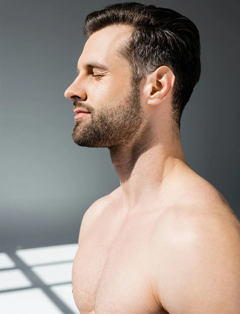 Profile view of a light-skinned, shirtless man with short, neatly styled dark hair and a short trimmed beard. His eyes are closed, and his expression is neutral and contemplative. His chest and shoulders show smooth, light skin with fine chest hair. The muted gray background features shadows cast by a light source, highlighting the contours of his face and body. - Renuvion for Men in Phoenix, AZ