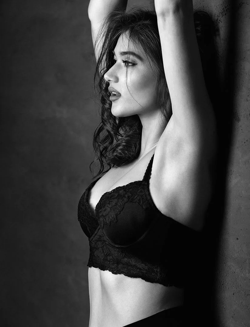 A dramatic, high-contrast, black and white profile photograph of a woman with long, dark, wavy hair, looking to the left. She is wearing a lace-trimmed black bralette, and her arms are raised above her head against a dark, textured wall. - Renuvion in Phoenix, AZ