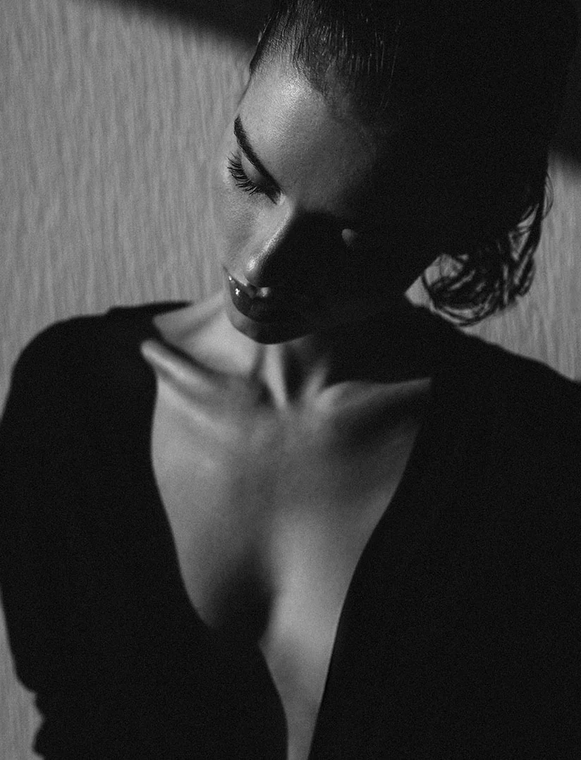 A moody, black and white close-up portrait of a woman looking down. She has slicked-back hair and is wearing a deeply plunging dark top that highlights her décolletage and collarbones. The lighting is dramatic, with deep shadows and a textured background wall. - Tuberous Breast Reduction in Phoenix, AZ